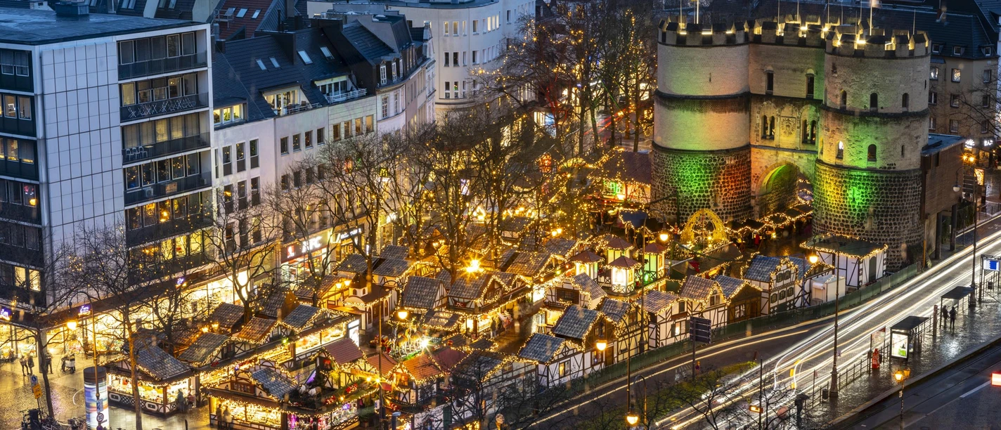 Christmas market on Rudolfplatz in Cologne with illuminated Hahnentor at dusk.