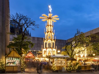 Adventsdorf am Kölner Dom Weihnachtspyramide mit Lichtern erhellt den Abend, umgeben von Marktständen und Besucher*innen.Christmas pyramid with lights lights up the evening, surrounded by market stalls and visitors.