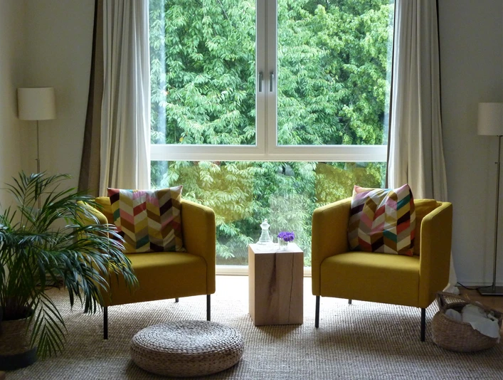 Ferienwohnung Zeitraum Zwei gelbe Sessel mit bunten Kissen stehen vor großen Fenstern, dahinter eine grüne Baumkulisse.