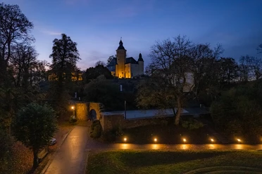 Nachtaufnahme der Burg Nümbrecht in herbstlicher Umgebung, stimmungsvoll beleuchtet, mit dunklem Himmel.