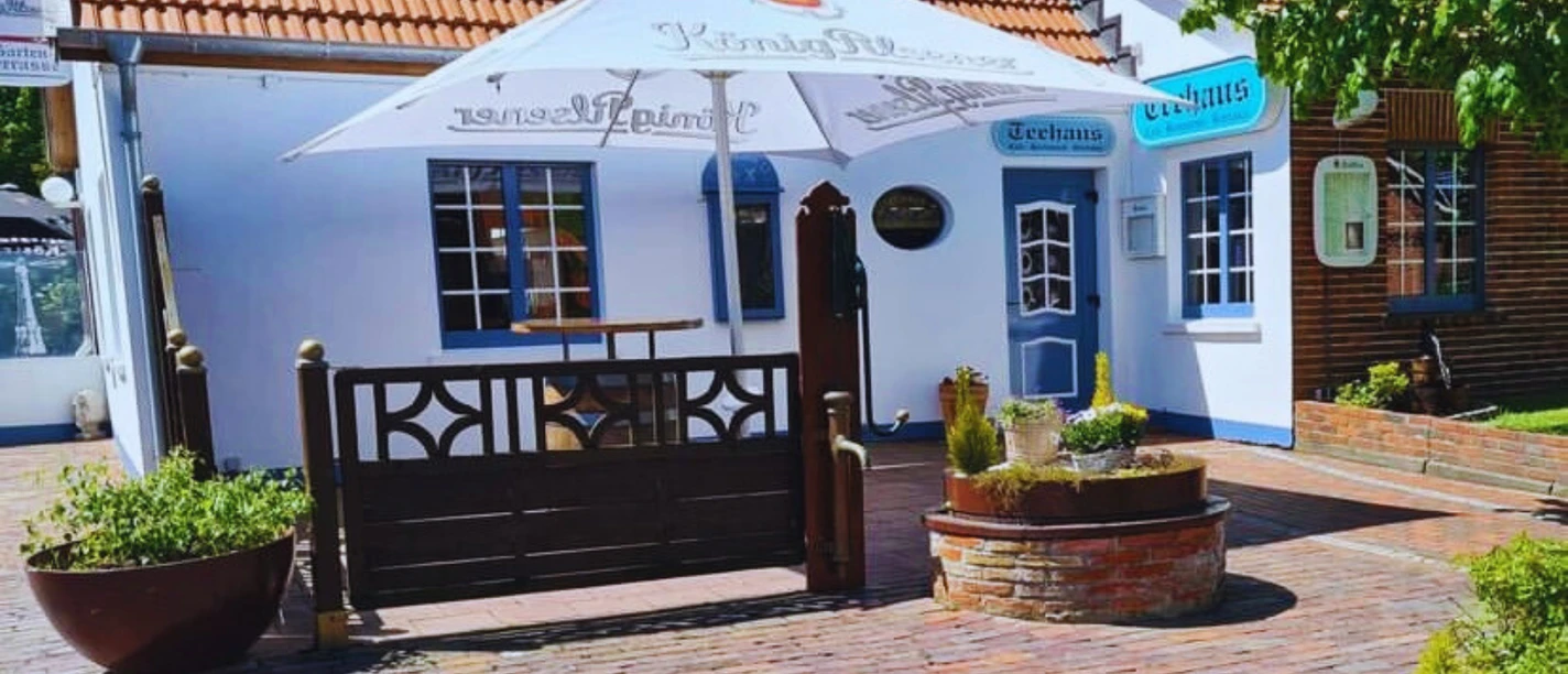The picture shows a charming white facade of a small tea house on the island of Borkum, adorned with blue shutters and surrounded by blooming plants. A rustic wooden sign reading 'Tea House' and an inviting sun umbrella create a relaxed and welcoming atmosphere. Numerous plant arrangements frame the cozy entrance area.