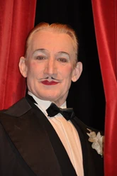 Robert Kreis.jpg Das Bild zeigt den Entertainer Robert Kreis in einem klassischen schwarzen Smoking mit Fliege vor einem roten Samtvorhang, während er lächelt.