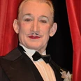 Robert Kreis.jpg Das Bild zeigt den Entertainer Robert Kreis in einem klassischen schwarzen Smoking mit Fliege vor einem roten Samtvorhang, während er lächelt.