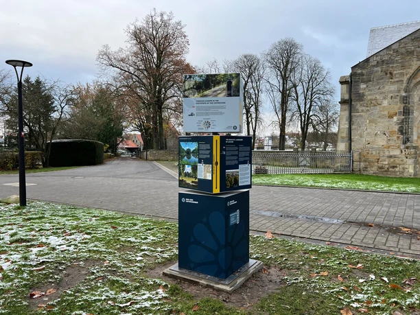 Informatiezuil in het groen voor het klooster van Loccum, geflankeerd door bomen en een pad op de achtergrond.
