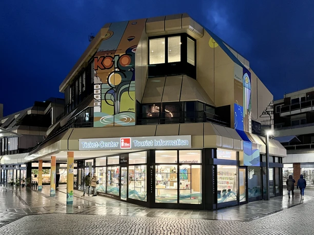 Tourist Information Paderborn, Außenansicht Tourist Information Paderborn mit leuchtendem Eingang in moderner Architektur bei Dämmerung.