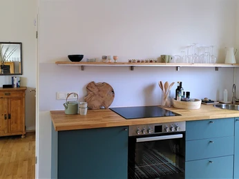 Ferienwohnung Zeitraum Moderne Küche mit blauen Unterschränken, Holzarbeitsplatte, Herd und offenen Regalen an der Wand.