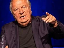 Ein älterer Mann in einem dunkelblauen Anzug spricht gestikulierend vor einem blauen Hintergrund.