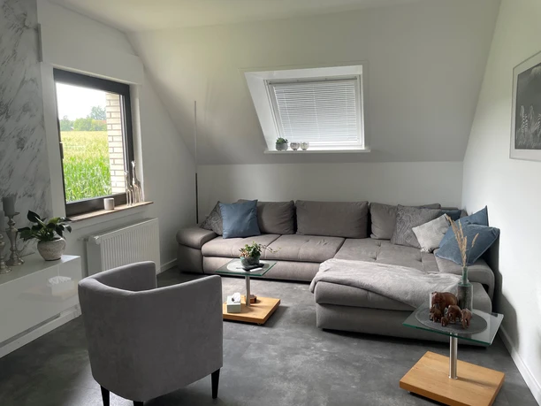 Wohnzimmer Wohnzimmer mit grauem Sofa, Sessel, Couchtischen; durch Fenster Blick auf ein grünes Feld.
