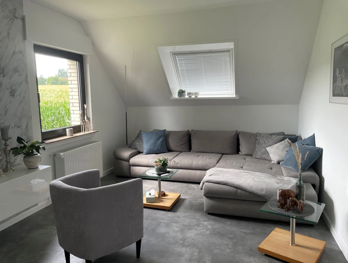 Wohnzimmer Wohnzimmer mit grauem Sofa, Sessel, Couchtischen; durch Fenster Blick auf ein grünes Feld.
