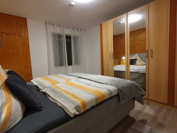 Schlafzimmer NEU Gemütliches Schlafzimmer mit Doppelbett, Kopfkissen in Grautönen, Holzmöbel und Fenster mit Vorhang.