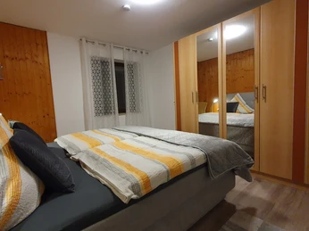 Schlafzimmer Gemütliches Schlafzimmer mit Doppelbett, Kopfkissen in Grautönen, Holzmöbel und Fenster mit Vorhang.