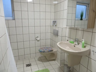 Badezimmer NEU Modernes, weiß gefliestes Badezimmer mit WC, Waschbecken, Spiegel und kleinen grünen Akzenten.