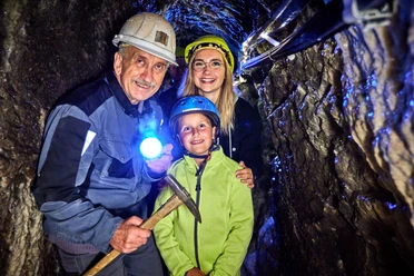 Kinderführung im Marie Louise Stolln Drei Personen mit Helmen im dunklen Bergwerksstollen, eine trägt eine Taschenlampe und lächelt.