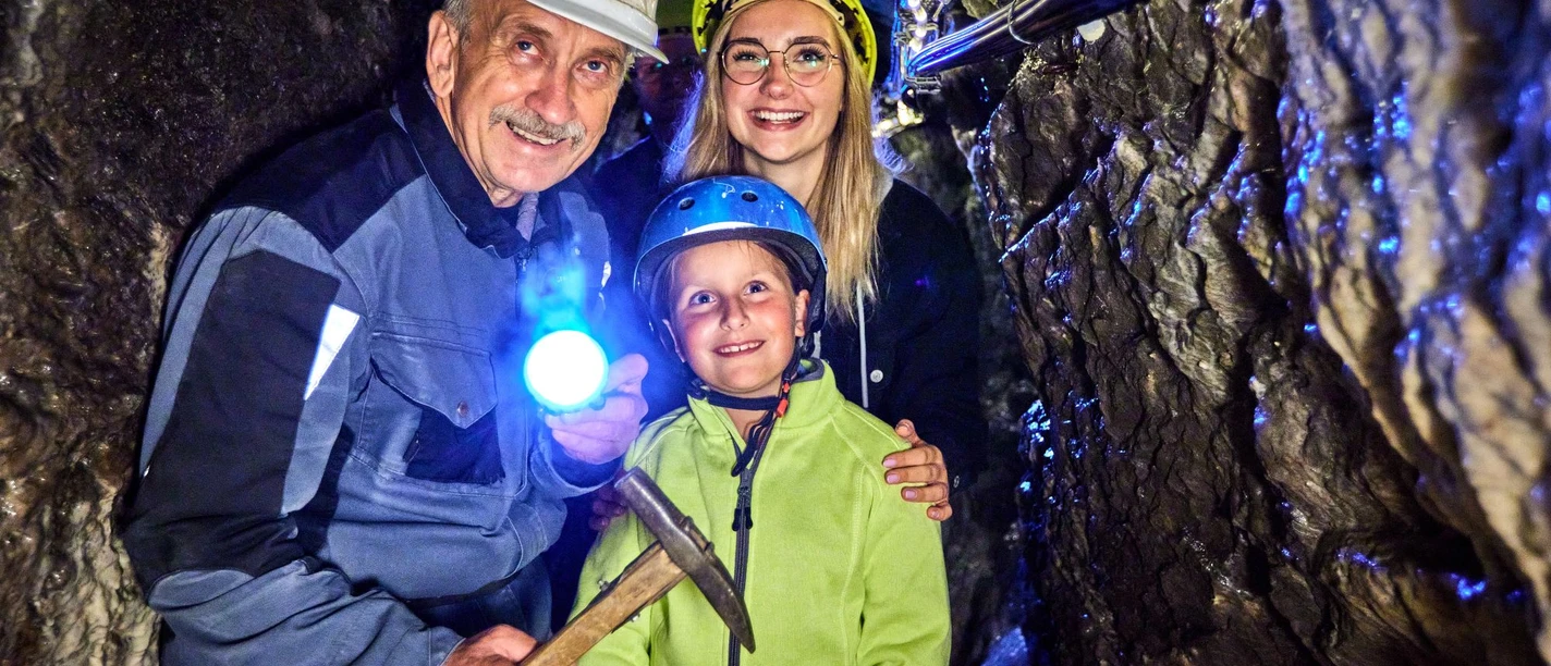 Kinderführung im Marie Louise Stolln Drei Personen mit Helmen im dunklen Bergwerksstollen, eine trägt eine Taschenlampe und lächelt.