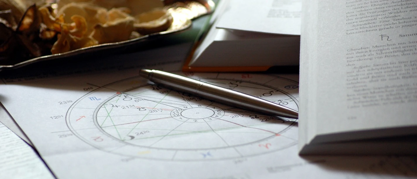 Astrologie Ein Horoskop und ein Stift liegen auf astrologischen Notizen zwischen geöffneten Büchern.
