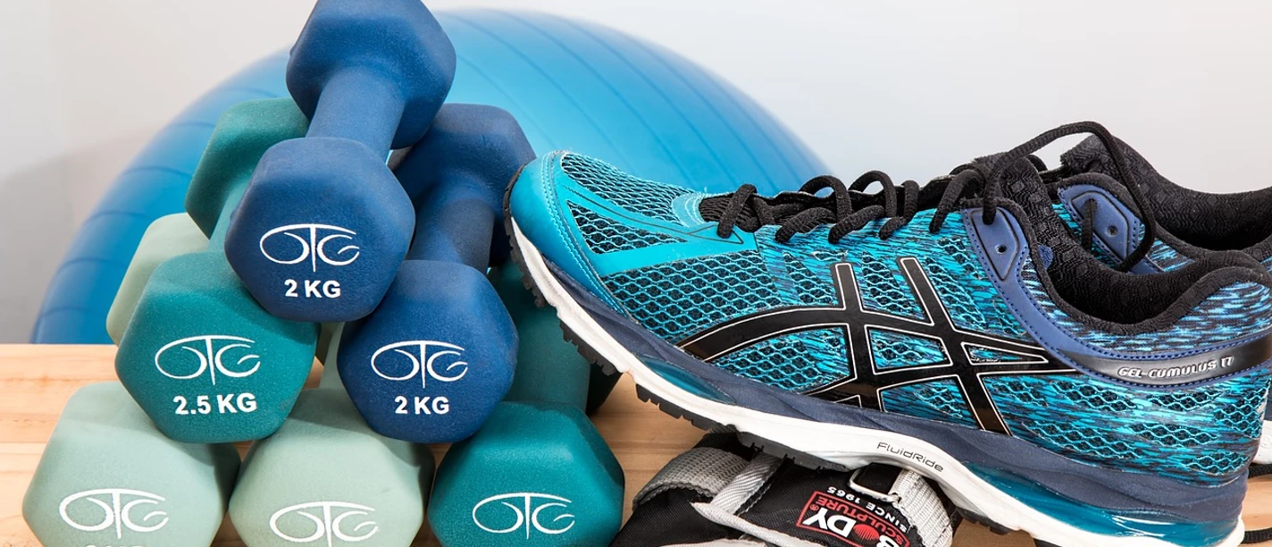 Bewegungskurs Hantelset, Turnschuhe, Fitnesshandschuhe und ein Gymnastikball vor hellem Hintergrund.