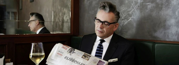Ein Mann in schwarzem Anzug mit Krawatte liest in einer Zeitung, ein Weißweinglas steht neben ihm.