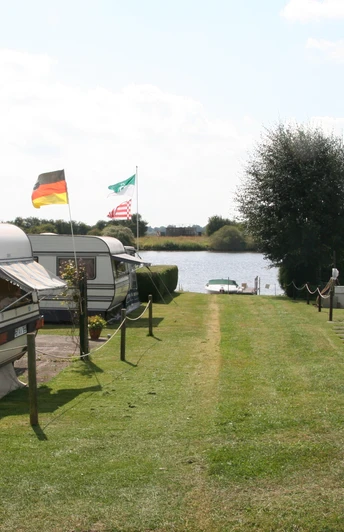 Ruhiger Campingplatz in Wassernähe mit Wohnwagen und Fahnen im Sommer, umgeben von grüner Natur.