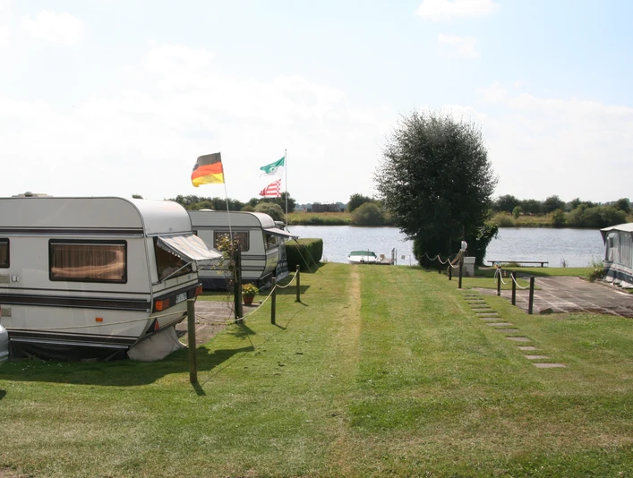 Ruhiger Campingplatz in Wassernähe mit Wohnwagen und Fahnen im Sommer, umgeben von grüner Natur.Quiet campsite near the water with caravans and flags in summer, surrounded by green nature.Rolig campingplads nær vandet med campingvogne og flag om sommeren, omgivet af grøn natur.Rustige camping aan het water met caravans en vlaggen in de zomer, omgeven door groene natuur.