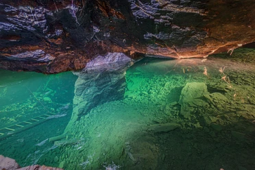 Underwater shot of a cave with greenish shimmering water, rocks and a ladder at the bottom, illuminated by warm light.