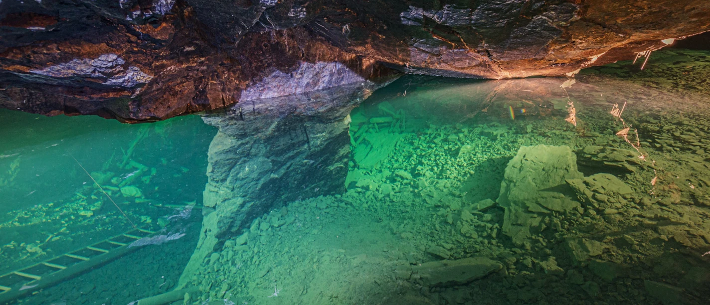 Underwater shot of a cave with greenish shimmering water, rocks and a ladder at the bottom, illuminated by warm light.