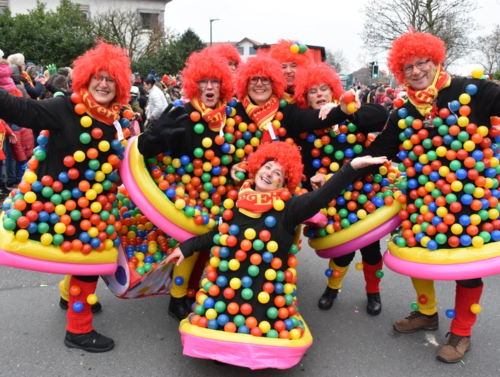 Menschen in bunten Kostümen mit roten Perücken und aufgeblasenen Ringen feiern ausgelassen Karneval.