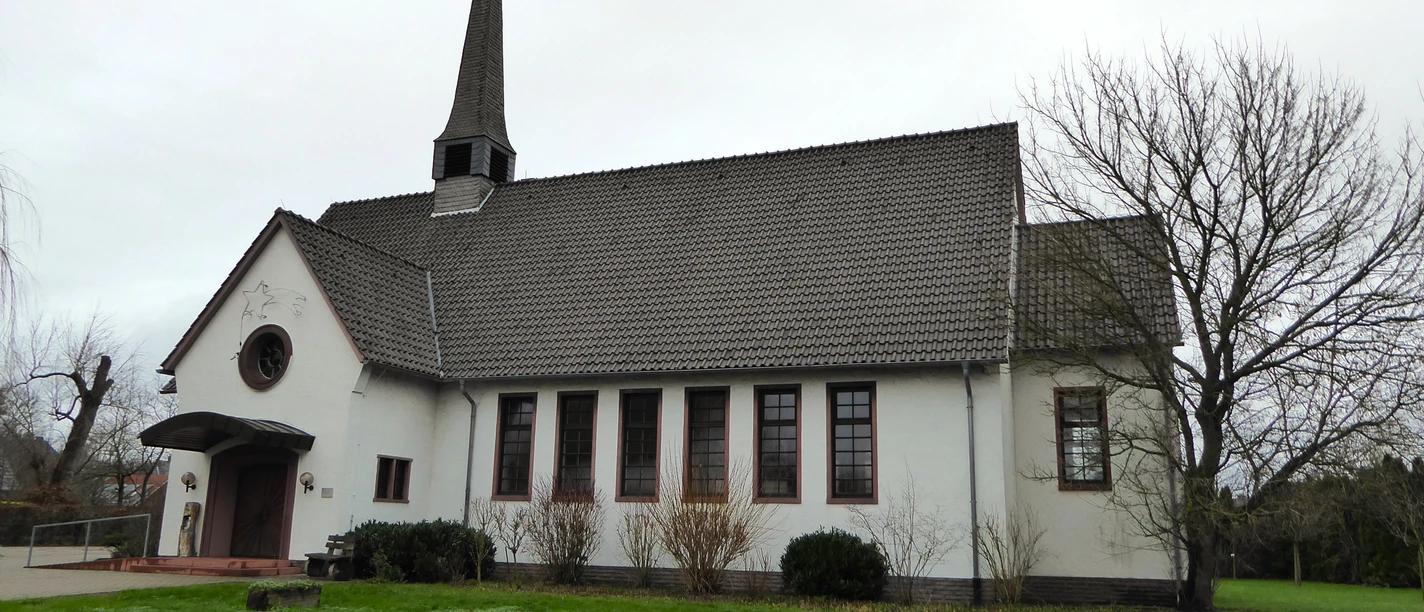 Kreuzkirche Herzebrock.JPG Ein kleines, weißes Kirchgebäude mit steilem, steinernem Turm und gepflegtem, grünem Rasen davor.