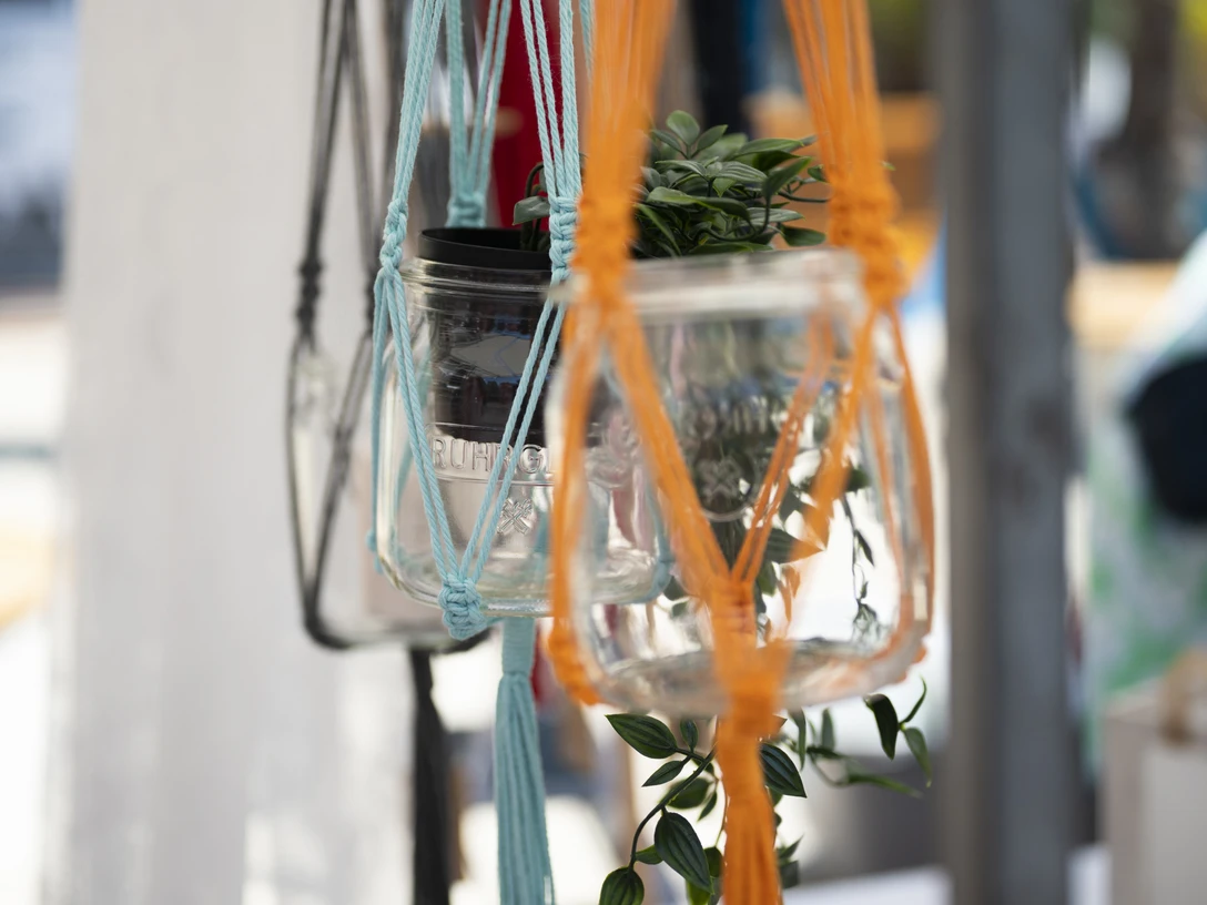 Herz und Hand.jpg Makramee-Blumenhalter mit bunten Schnüren und Glasgefäßen, darin kleine grüne Pflanze.