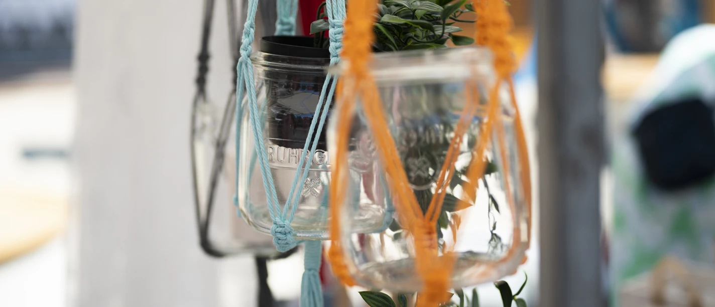 Herz und Hand.jpg Makramee-Blumenhalter mit bunten Schnüren und Glasgefäßen, darin kleine grüne Pflanze.