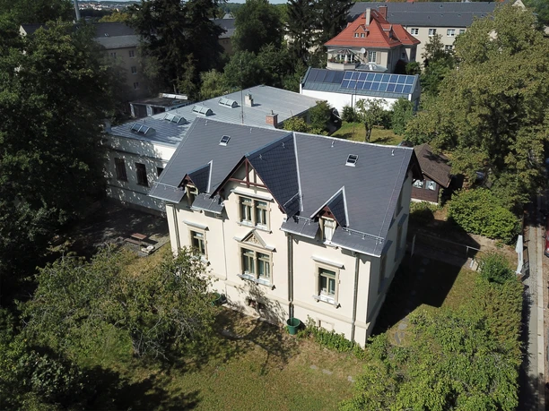 Schaustickerei Altes Gebäude mit grau-blauem Dach, umgeben von Bäumen und weiteren Gebäuden im Hintergrund.