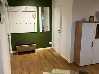 Garderobe.jpg Moderner Flur mit grüner Akzentwand, Kleiderhaken, Sitzbank und Spiegel, Holzfußboden.