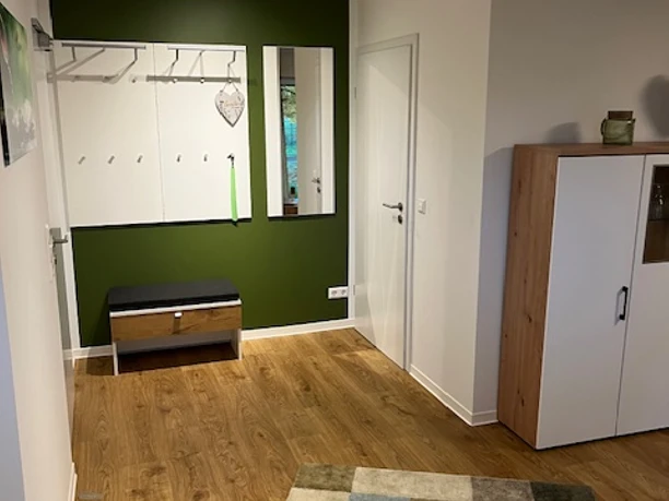 Garderobe.jpg Moderner Flur mit grüner Akzentwand, Kleiderhaken, Sitzbank und Spiegel, Holzfußboden.