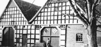 bildanzeige-1.jpg Fachwerkhof mit drei Gebäuden, Giebelseiten, eine Person am Tor, Baum im Vordergrund. 20. Jahrhundert.