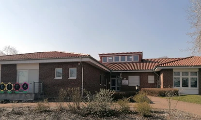 kindergarten_tausendfueßler_ahlsen_01.jpg Barrierefreier Eingangsbereich eines einstöckigen Ziegelgebäudes mit rotem Ziegeldach unter blauem Himmel.