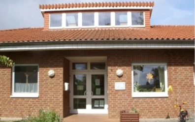 kindergarten_tausendfueßler_ahlsen.jpg Backsteinbau eines Kindergartens mit Fenstergiebel und dekorierter Eingangstür, umrahmt von Pflanzen.