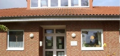 kindergarten_tausendfueßler_ahlsen.jpg Backsteinbau eines Kindergartens mit Fenstergiebel und dekorierter Eingangstür, umrahmt von Pflanzen.