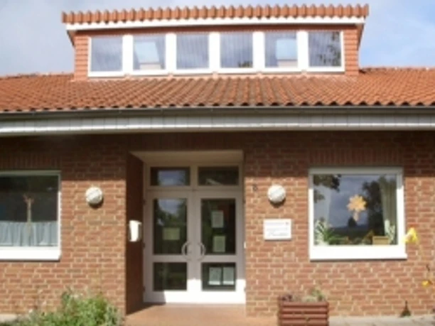 kindergarten_tausendfueßler_ahlsen.jpg Backsteinbau eines Kindergartens mit Fenstergiebel und dekorierter Eingangstür, umrahmt von Pflanzen.