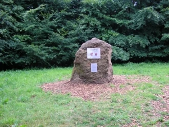 Ein markanter Stein steht auf einer von Bäumen umgebenen Lichtung und trägt historische Tafeln.