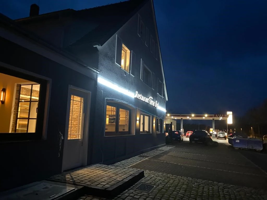 TimeOut Außenansicht Ein beleuchtetes Restaurant neben einer Tankstelle in der Abenddämmerung, mit parkenden Autos davor.