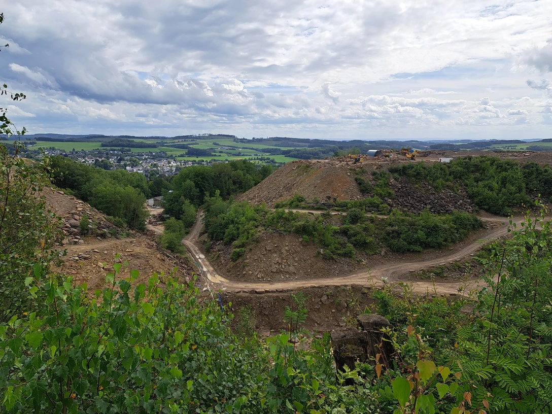 Steinbruch in Lindlar Steinbruch mit grünen Hügeln im Hintergrund, zahlreiche Baugeräte und wolkiger Himmel darüber.