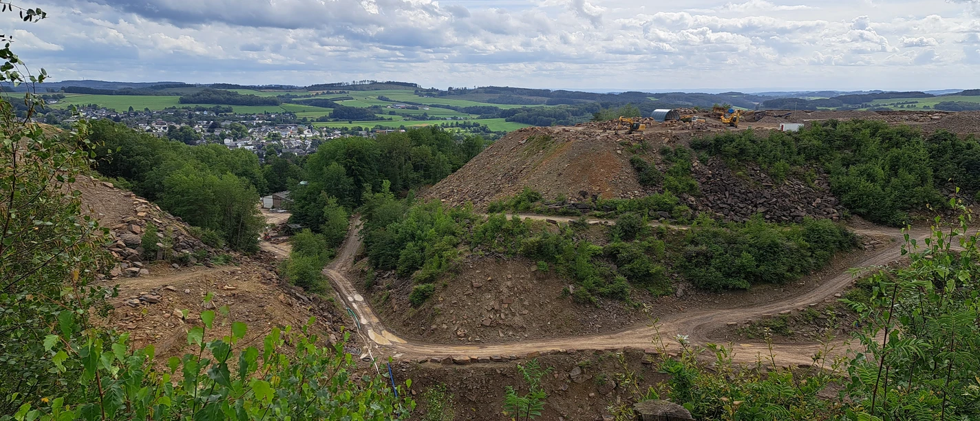 Steinbruch in Lindlar Steinbruch mit grünen Hügeln im Hintergrund, zahlreiche Baugeräte und wolkiger Himmel darüber.