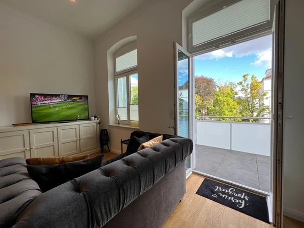 Wohnbereich Heller Wohnbereich mit Sofa, offenem Balkonblick auf grüne Bäume, TV auf Sideboard.