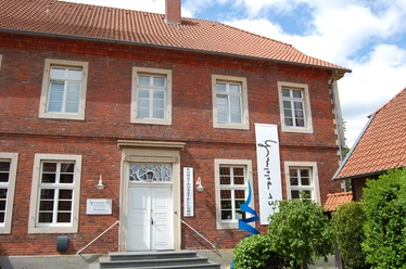 Galerie 13.jpg Historisches Gebäude aus rotem Backstein mit weißen Fenstern und Bannern am Eingang.