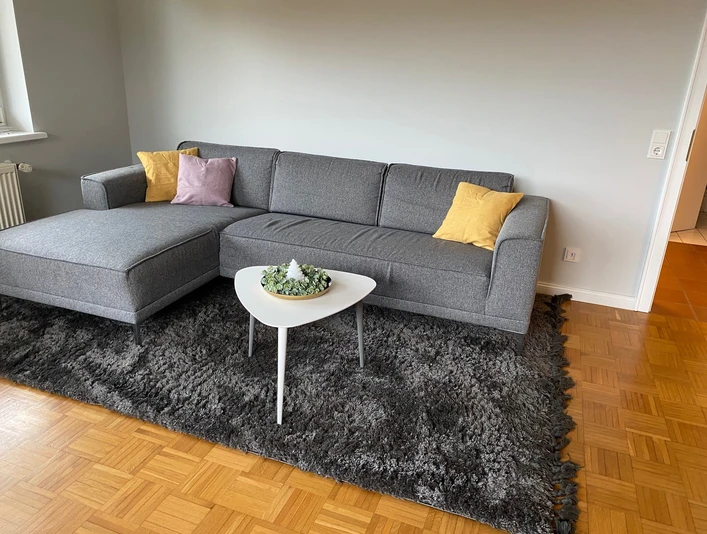 Ein modernes Wohnzimmer mit grauer Couch, bunten Kissen und einem weißen Couchtisch auf Teppich.
