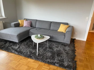 holsing.jpg Ein modernes Wohnzimmer mit grauer Couch, bunten Kissen und einem weißen Couchtisch auf Teppich.