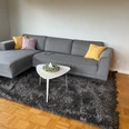 holsing.jpg Ein modernes Wohnzimmer mit grauer Couch, bunten Kissen und einem weißen Couchtisch auf Teppich.