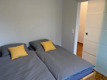 Zwei Einzelbetten mit grauer Bettwäsche und gelben Kissen in einem minimalistischen Schlafraum.