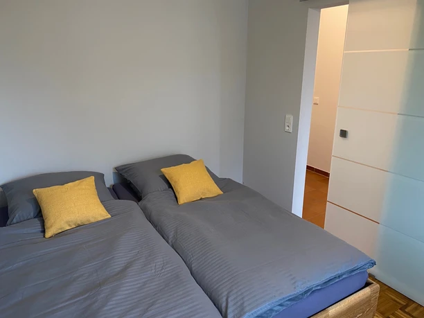 holsing4.jpg Zwei Einzelbetten mit grauer Bettwäsche und gelben Kissen in einem minimalistischen Schlafraum.