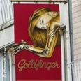 Ein auffälliges, rotes Schild mit einer goldenen Frau und dem Text "Goldfinger" in geschwungener Schrift.