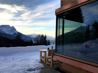 Tannenhütte Verschneite Terrasse mit Holzbank, Bergkulisse und Glasfront der Hütte im Abendlicht